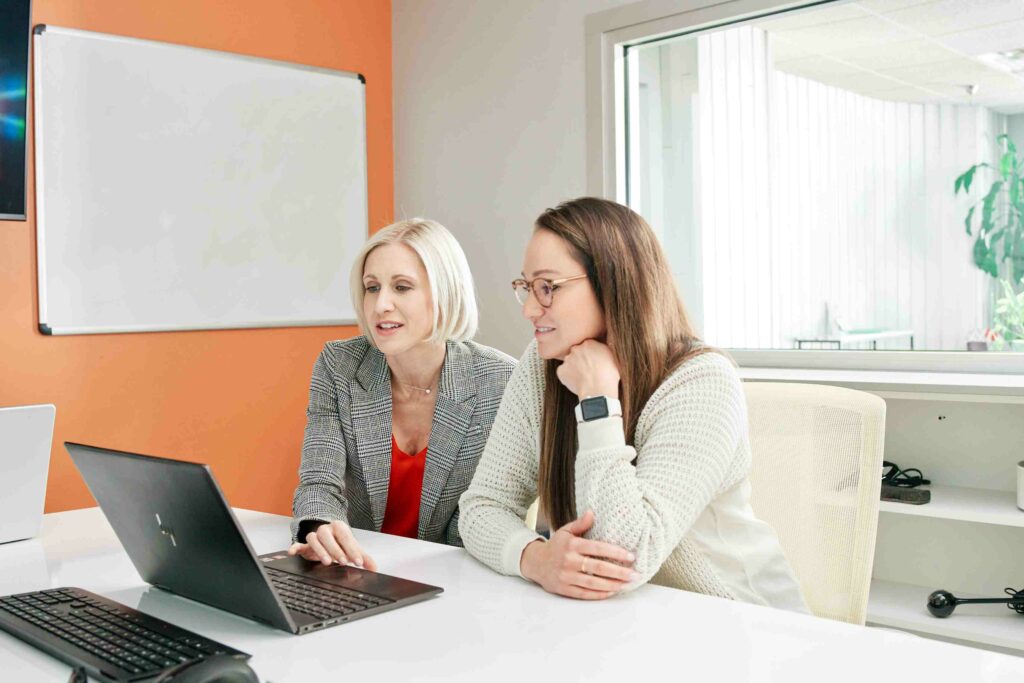 Two professional women evaluate data on a laptop in a brightly lit office.