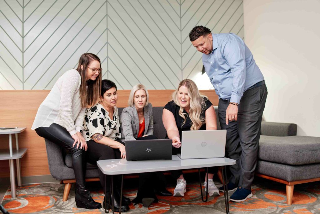 Five upbeat colleagues collaborate around a laptop amidst a vibrant office setting.
