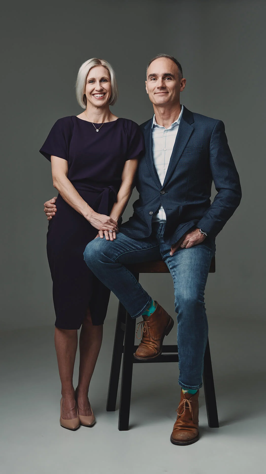 A professionally dressed, cheerful duo poses together against a gray backdrop.