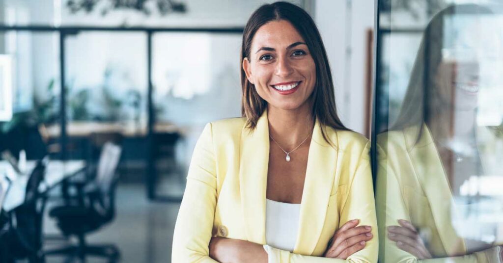Confident woman in yellow blazer stands by reflective surface in office.